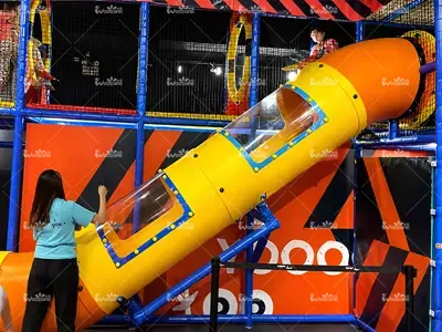 Bright yellow and orange enclosed tube slide in an indoor playground, with children playing inside the multi-level soft play structure