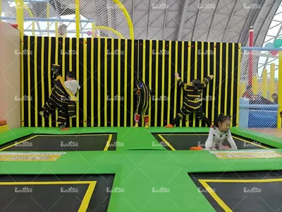 Children playing on black and yellow vertical sticky wall with green padded freestyle trampoline zone inside indoor trampoline park